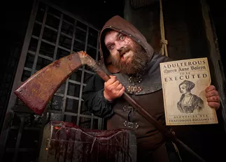 The Rotten Royals executioner is posing inside the London Dungeon with his axe and sign detailing the execution of Anne Boleyn. The London Dungeon is a popular tourist attraction in London that offers a variety of interactive experiences based on London's history.